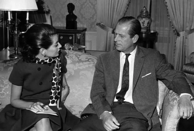 A woman and a man seated on a sofa in formal attire, engaging in conversation in an elegantly decorated room.