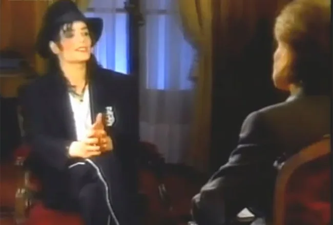 Two people in a formal interview setting, one in a black hat and suit, gesturing with hands in a warmly lit room.
