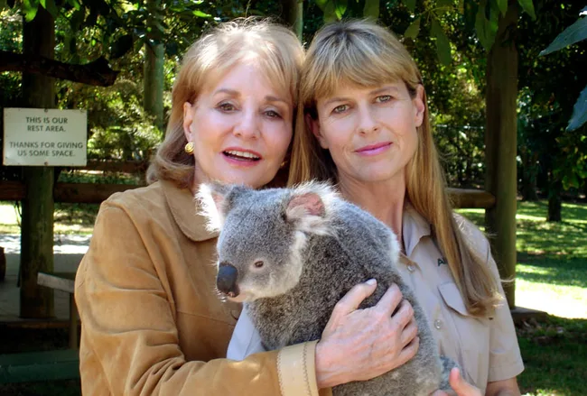 Two women smiling, one in a brown coat, the other in a uniform, holding a koala, standing in a green park setting.