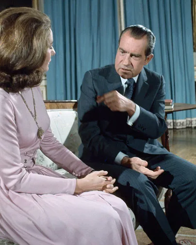 Man in a suit gestures while talking to a woman in a pink dress sitting on a floral couch indoors.