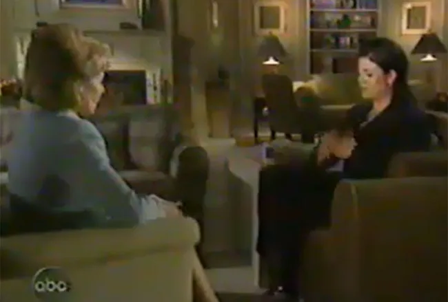 Two people seated in a living room setting having a conversation, with a fireplace and bookshelves in the background.