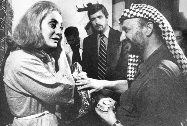A woman in a robe interacts with a man in a keffiyeh, while others look on in a room with patterned decor.