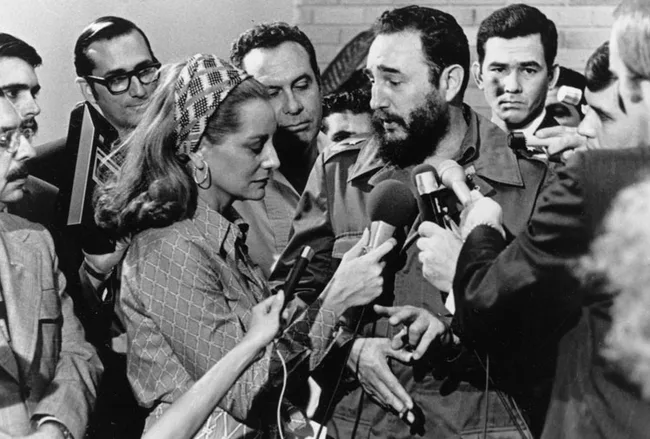 Group of journalists interviewing a bearded man in military attire, speaking into multiple microphones.