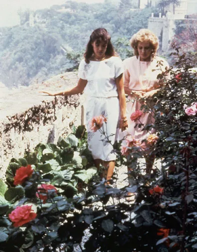 Two women walking in a rose garden, one in a white dress and one in pink, with a scenic view in the background.