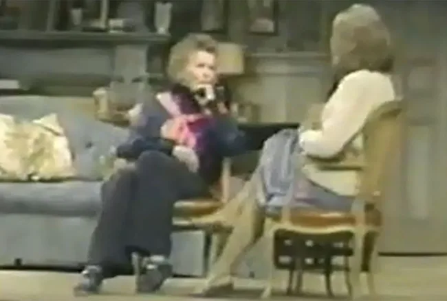 Two women sitting and talking in a cozy living room setting, with one on a sofa and the other on a chair.