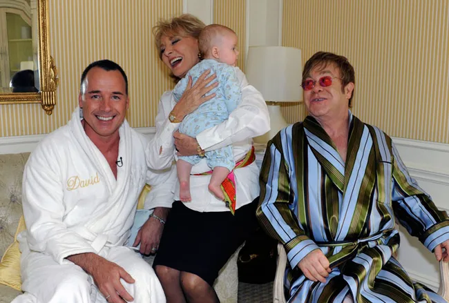 Man in robe, woman holding baby, and another man in striped robe sitting in a room with patterned wallpaper.