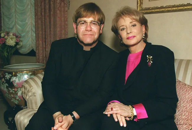 Two people sitting on a sofa, one wearing glasses and black attire, the other in pink and black, in a warmly decorated room.