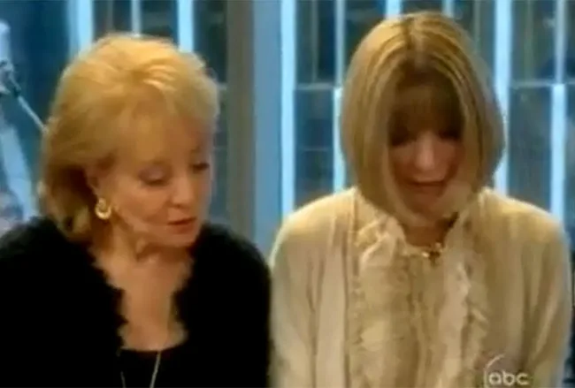 Two women conversing closely indoors, one with short blonde hair wearing earrings, the other with bob-cut hair and a white top.