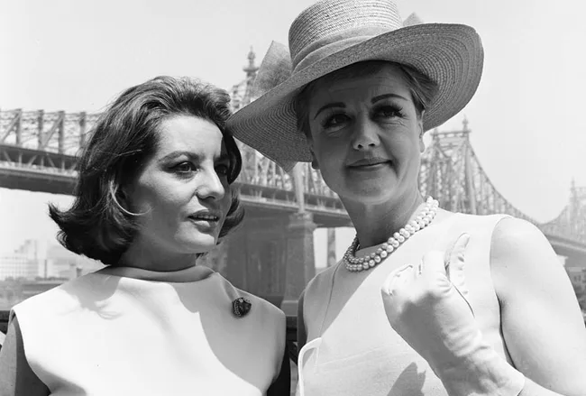 Two women in 1960s attire, one wearing a hat and pearls, standing in front of a bridge.