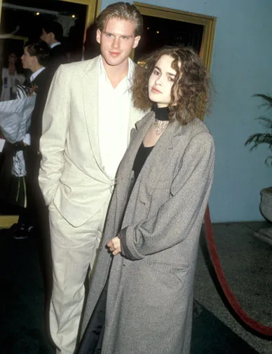 A man in a light suit and a woman in a long coat pose together on a red carpet.