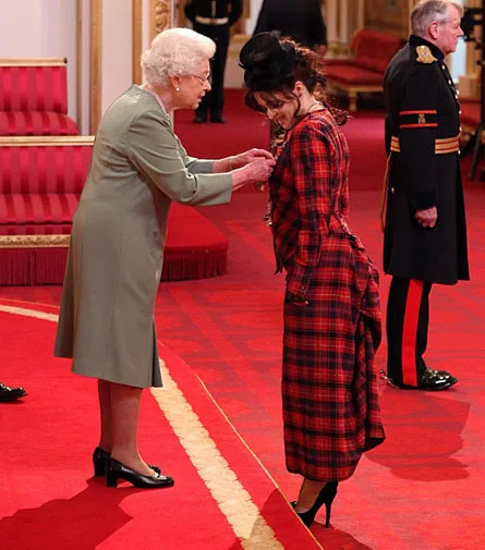 A woman in a green outfit pins a medal on another woman in a red plaid dress inside a formal room with red decor.