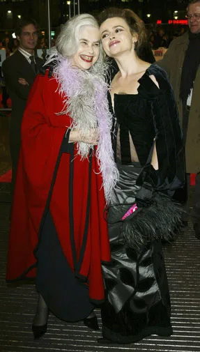 Two women pose together; one in a red coat with feathers, the other in a black gown holding a clutch.