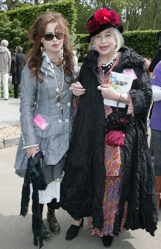 Two women in stylish, eclectic outfits and sunglasses stand together outdoors, one in a coat, the other in a floral dress.