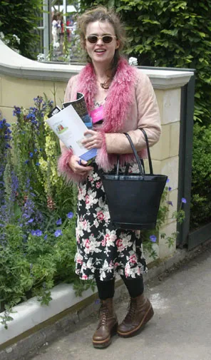Woman in floral dress with pink fur collar, holding documents and a black bag, standing outdoors by a garden.