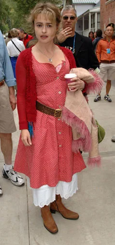 A woman in a red polka dot dress and boots holds a coffee cup, standing on a street with people in the background.