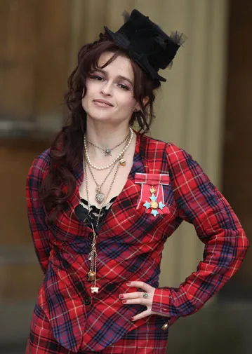 Helena Bonham Carter in a red plaid jacket with medals and a small black hat, posing outdoors.