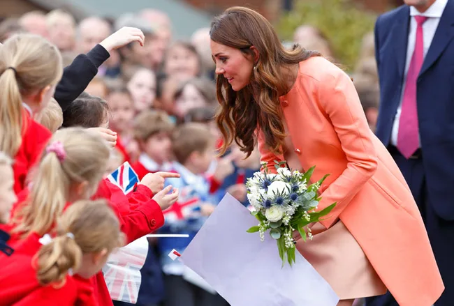 A woman in an orange coat greets children and receives flowers during a public event.
