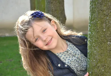 Young girl smiling, peeking from behind a tree, wearing a blue flower headband and patterned shirt, outdoors.