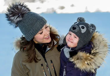 Mother and child in winter attire smiling in snowy landscape, both wearing animal-themed hats.