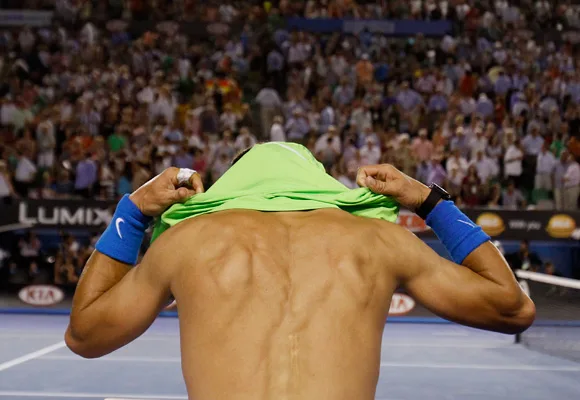 "Shirtless tennis player pulls green jersey over head on court, with crowd cheering in background."