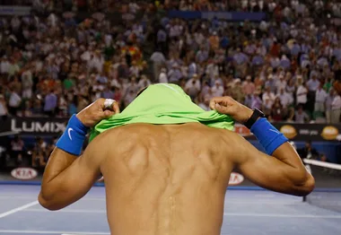 "Shirtless tennis player pulls green jersey over head on court, with crowd cheering in background."