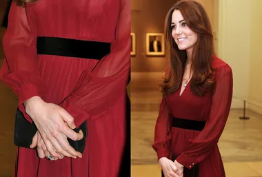 A woman in a red dress with a black belt and subtle baby bump stands smiling, hands gently clasped.