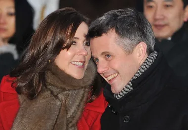 Couple smiling warmly at each other, dressed in winter coats with fur details, surrounded by blurred onlookers.