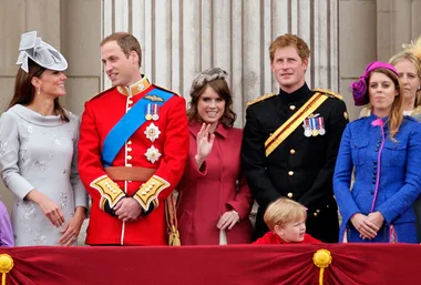 A group of royals, including adults in formal attire and a child, stand on a balcony at a public celebration.