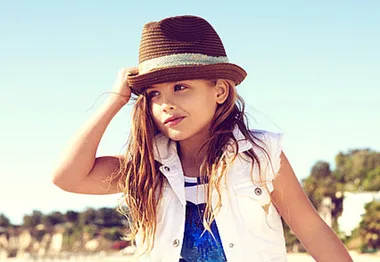 Young girl wearing a brown hat and white vest, standing outdoors with a thoughtful expression.