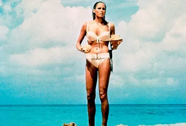 A woman in a white bikini stands on a beach with a shell in hand, a scene from a classic film against a blue sky and ocean.