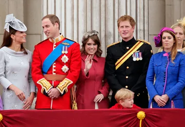 A group of royals on a balcony during a formal event, dressed in ceremonial attire, with one child peeking over the rail.