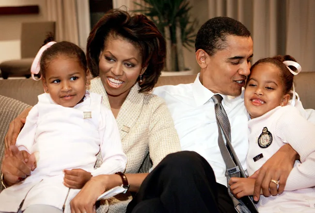 A family of four sitting together on a couch, with parents and two young children smiling and embracing each other.