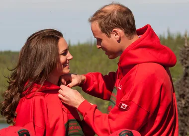 A couple in red hoodies smile warmly at each other outdoors, with trees in the background.