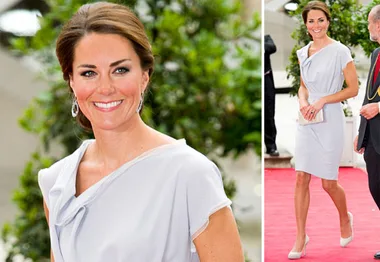 A woman in a light grey dress on a red carpet, smiling and holding a clutch, with greenery in the background.