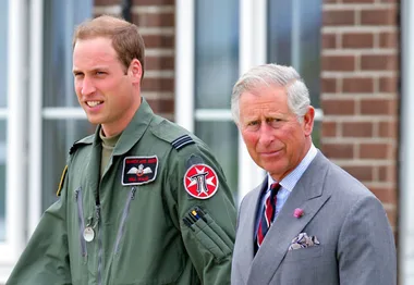 Two men walking outside; one in a military uniform, the other in a suit, against a brick and windowed background.