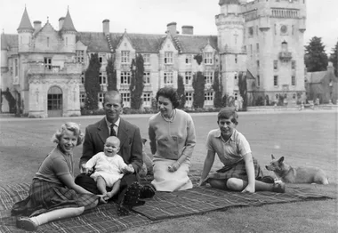 A family sitting on a blanket with a corgi outside a large castle-like building.