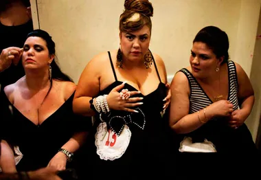 Three women in black dresses, one with a contestant number, preparing for a pageant in a backstage area.