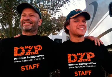 Two men wearing Dartmoor Zoological Park staff shirts and hats, smiling with arms around each other outside near trees.
