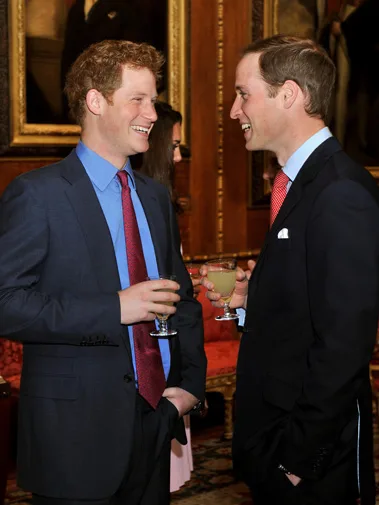 Two men in suits having a conversation while holding drinks, set in an elegant room with framed portraits on the walls.