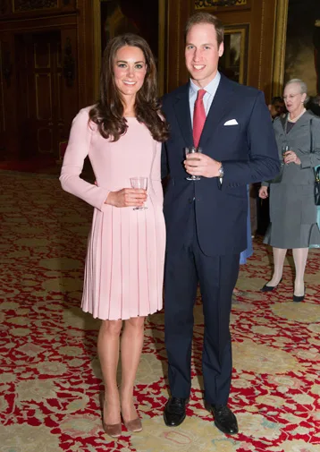 A couple posing together, the woman in a pink dress and the man in a suit, holding drinks in an elegant room.