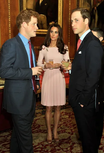 Three individuals in formal attire conversing at an indoor event with ornate decor, holding drinks.