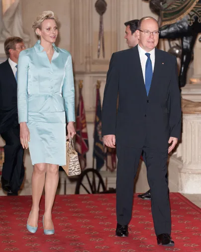 A man in a suit and a woman in a light blue outfit walk on a red carpet in a grand hall with flags in the background.