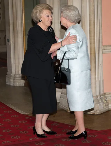 Two women dressed elegantly, greeting each other warmly in a formal setting with red carpeted flooring.