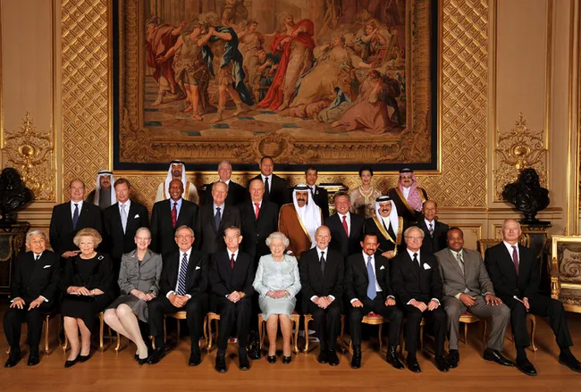 Group of dignitaries in formal attire seated and standing in an ornate room with a large tapestry behind them.
