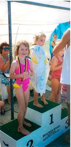 Children stand on a podium at a swimming event, with medals and towels; girl on podium step 2 is in a pink swimsuit.