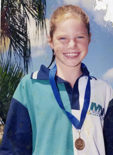 A young girl smiling outdoors, wearing a school sports uniform and a medal around her neck.