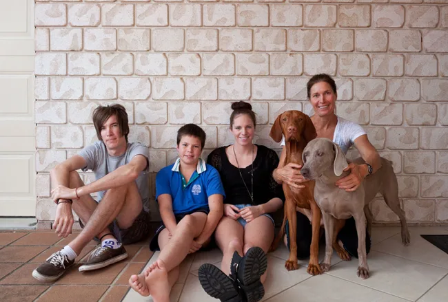 Family with three children and two dogs sitting against a brick wall, smiling.