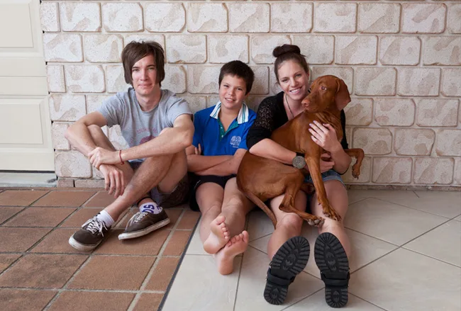 Three people sitting on a tiled floor against a brick wall, smiling, with a brown dog.