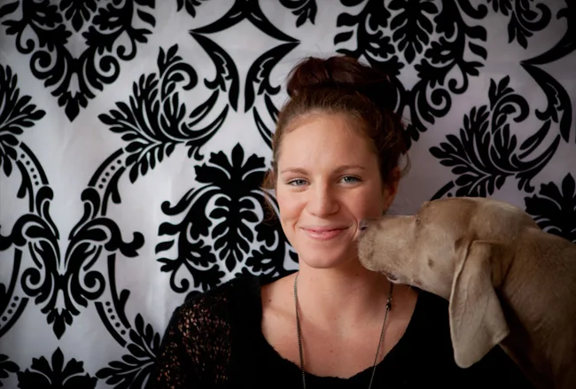 Young woman smiles as a dog affectionately nuzzles her cheek against a decorative black and white patterned background.