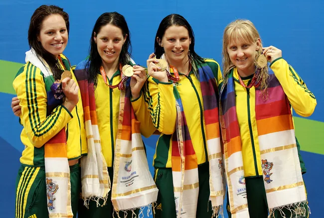 Australian women's swimming team smiling and holding medals at the 2010 Commonwealth Games, wearing yellow and green tracksuits.